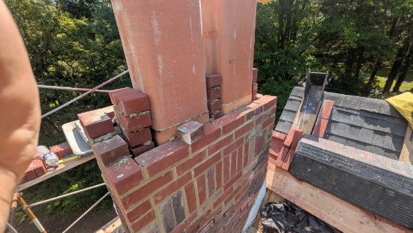 Chimney top rebuild in progress with brick being laid around the two clay flue liners on scaffolding above the roof, York County PA